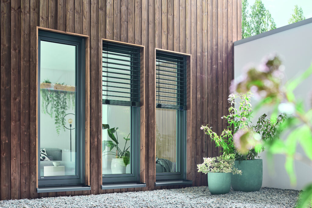 alt="Moderne Kunststoff-Alu-Fenster mit schlanken Rahmen und außenliegendem Sonnenschutz an einer Fassade mit Holzverkleidung – energieeffizient, pflegeleicht und ideal für zeitgemäßen Wohnkomfort"